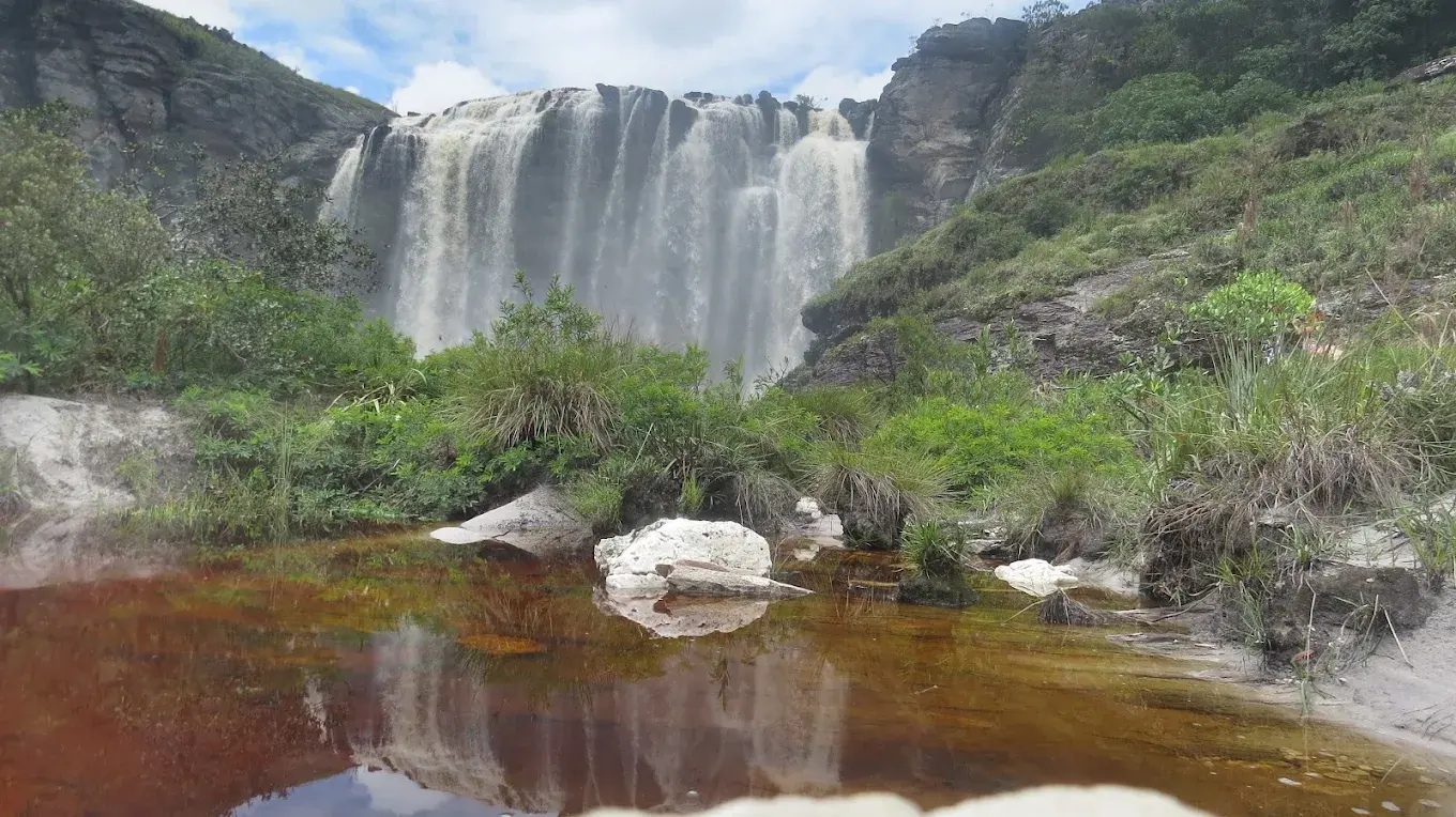 Cachoeira ampla caindo sobre paredão de pedra até um poço com água avermelhada, em Lapinha da Serra