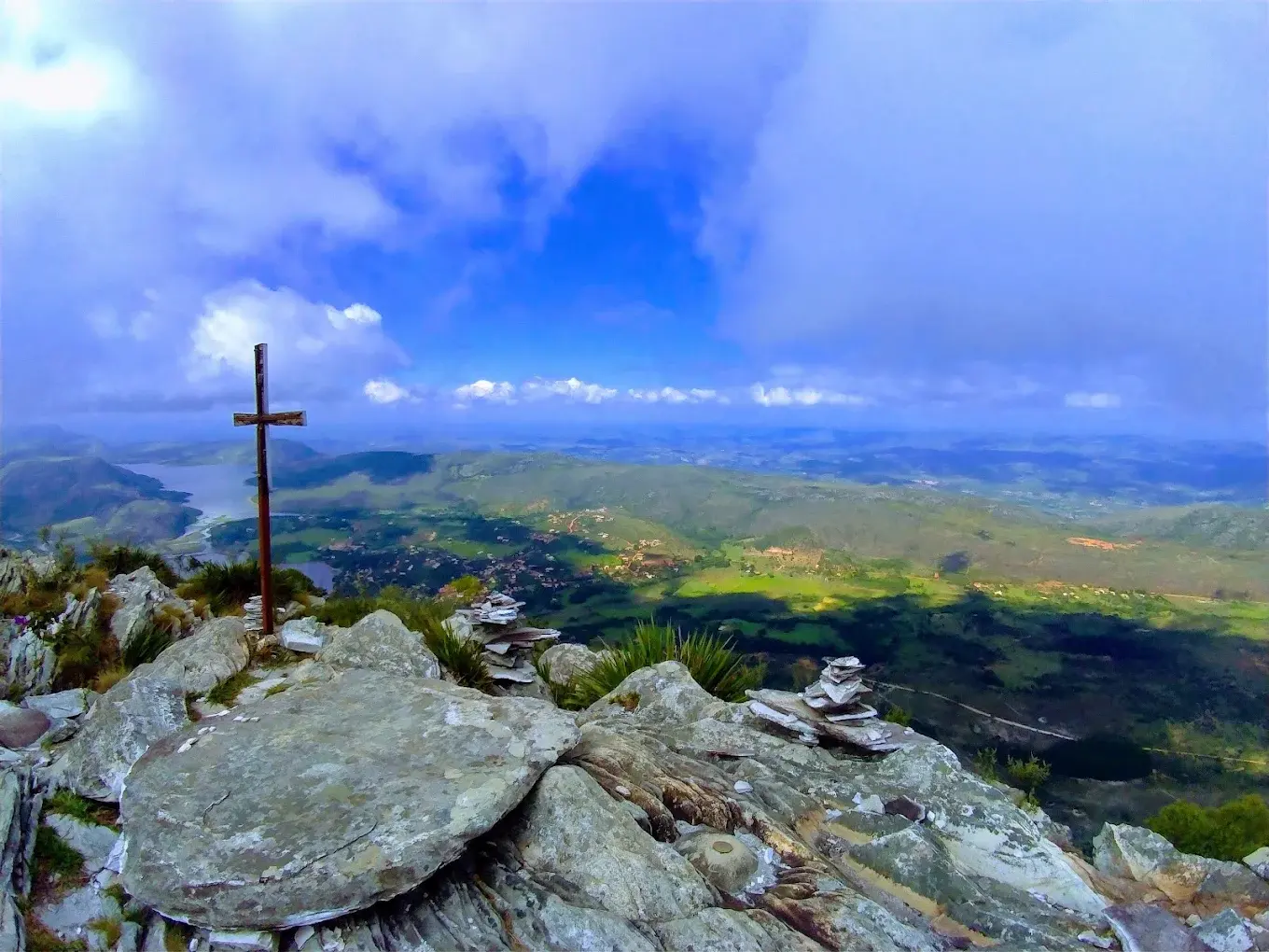 Cruzeiro no cume do Pico da Lapinha em dia de céu aberto, com o vilarejo e a represa ao fundo